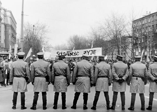 Niezależna manifestacja pierwszomajowa. Na pierwszym planie kordon milicjantów, blokujący przemarsz pochodu, w tle manifestanci z hasłem Solidarność żyje.